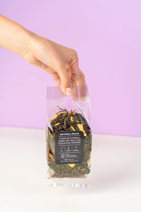 Mano sostiene una bolsa de té Natural Peace sobre fondo lila.