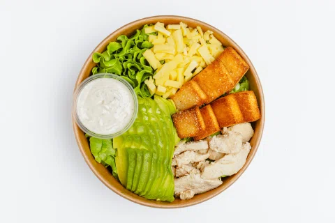 Bowl con pollo, palta, hojas verdes, queso y salsa sobre fondo blanco.