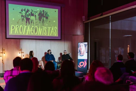 Panel de conversación frente al público en un salón con iluminación rosada.