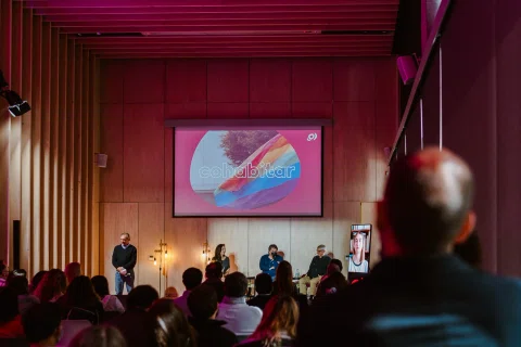 Público observa una charla en un salón iluminado en tonos rosados.
