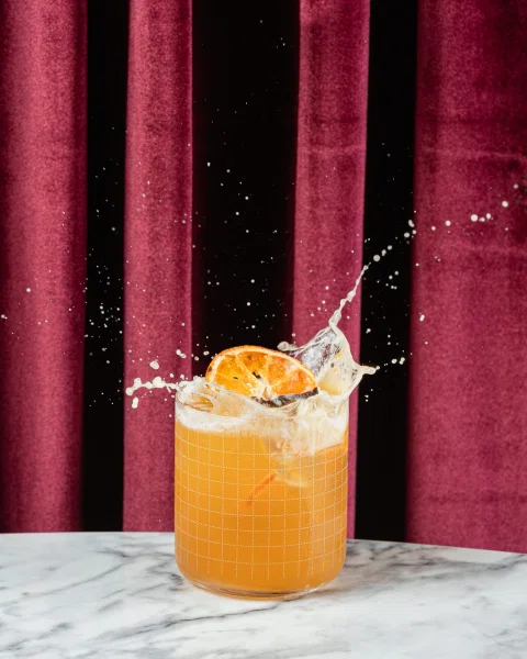 Cóctel naranja con hielo, rodaja de cítrico y una salpicadura frente a fondo rojo.