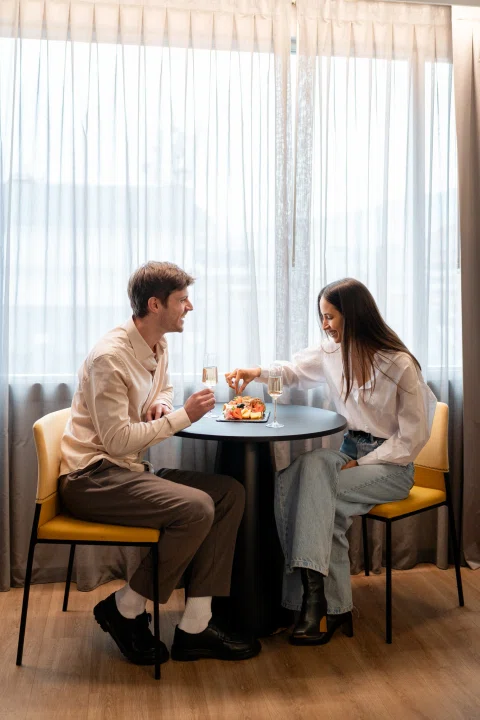 Dos personas compartiendo comida en mesa junto a ventana.