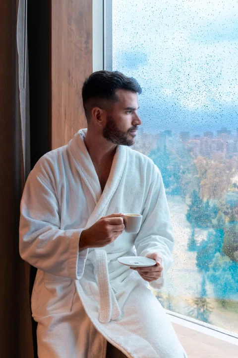 Hombre con bata tomando café frente a ventana.