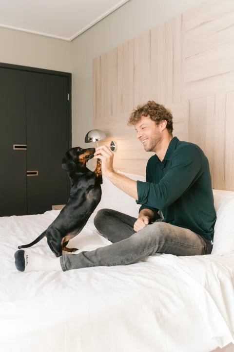 Hombre jugando con perro sobre cama de hotel.
