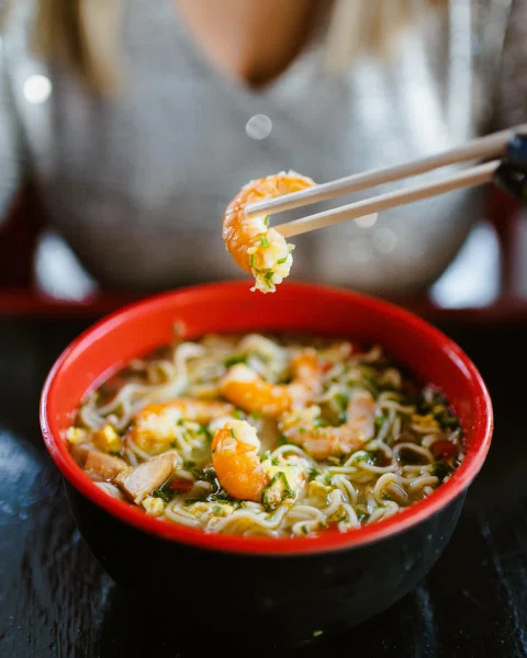 Mano levantando camarón desde bowl de ramen.