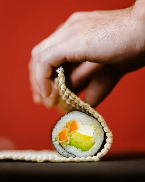 Mano sosteniendo una pieza de sushi frente a fondo rojo.