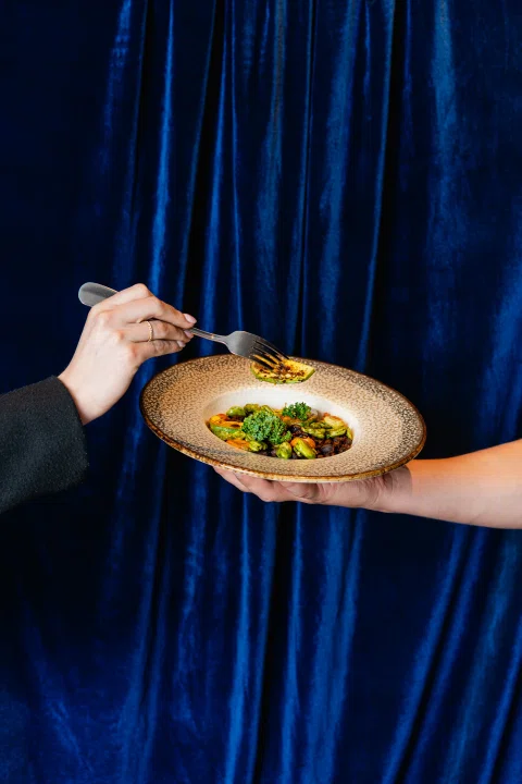 Mano sosteniendo plato gastronómico frente a cortina azul.