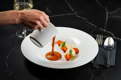 Mano vertiendo salsa sobre entrada gastronómica.