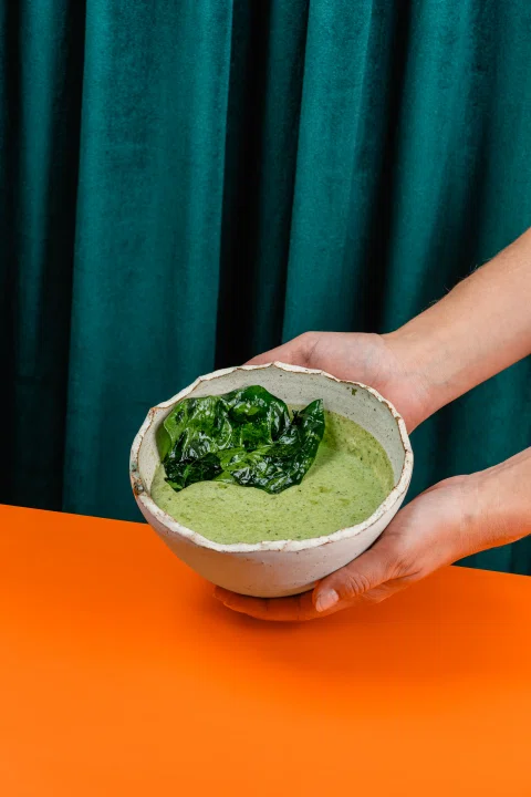 Manos sosteniendo bowl con preparación verde sobre fondo naranja.