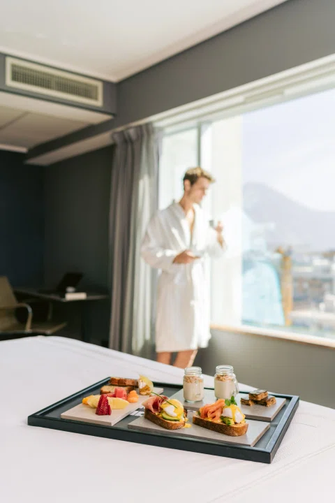 Mujer con bata junto a bandeja de desayuno y ventana.
