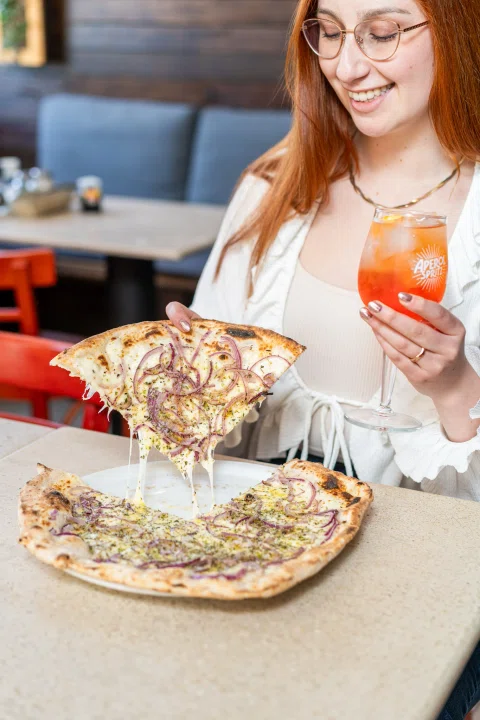 Mujer comiendo pizza acompañada de cóctel naranja.