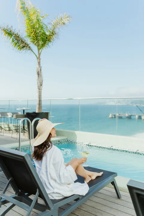 Mujer leyendo en reposera frente a piscina y mar.
