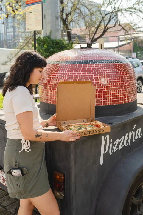 Mujer llevando caja de pizza frente al horno exterior.