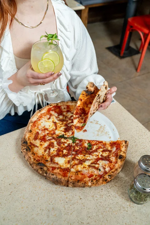 Mujer sirviendo una porción de pizza junto a cóctel.