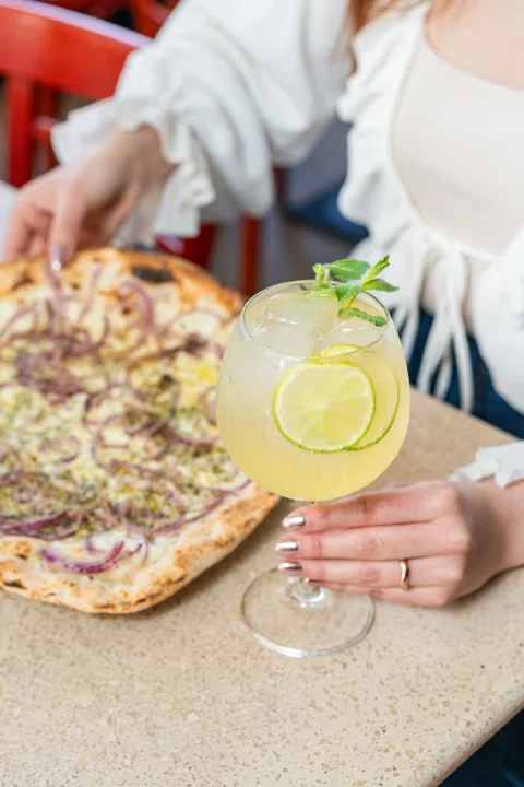 Persona con pizza y cóctel claro en mesa.