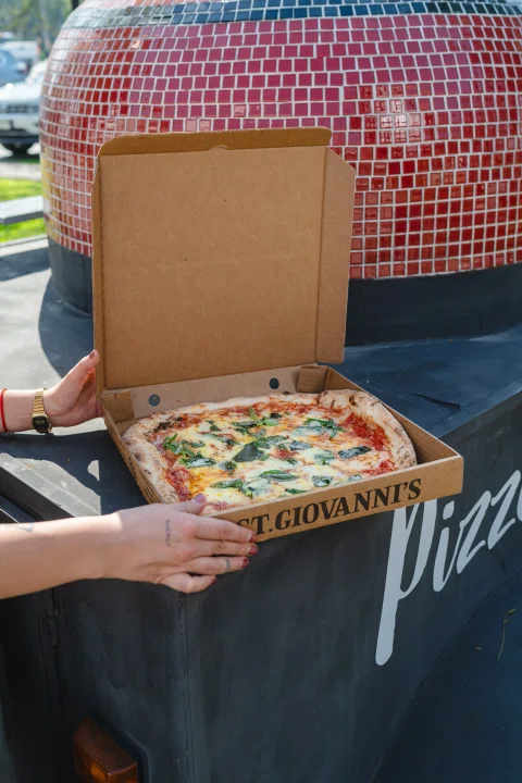 Persona sosteniendo caja abierta de pizza frente a horno.