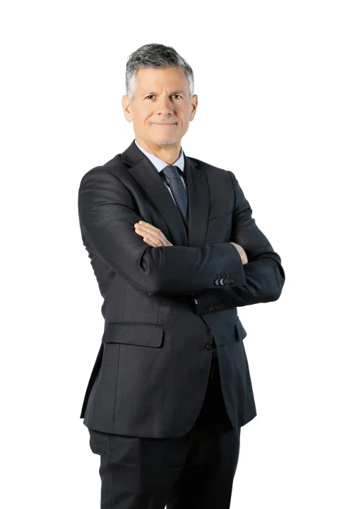 Retrato corporativo de un hombre con traje negro y brazos cruzados sobre fondo blanco.