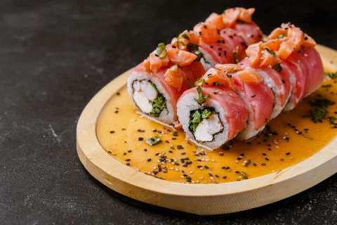 Roll de sushi cubierto con salmón y salsa sobre tabla.