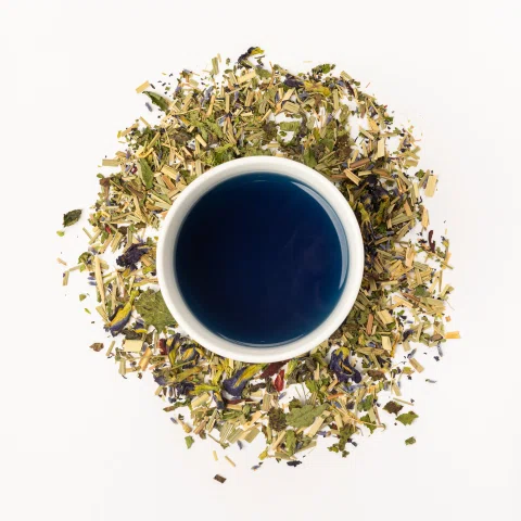 Taza azul con infusión oscura rodeada por hierbas.