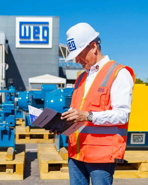 Técnico de WEG con casco y chaleco de seguridad revisa documentación junto a equipos industriales.