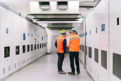 Dos técnicos recorren una sala industrial entre filas de tableros eléctricos.