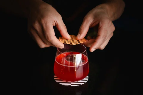 Manos sostienen una galleta sobre un cóctel rojo en una composición de fondo oscuro.